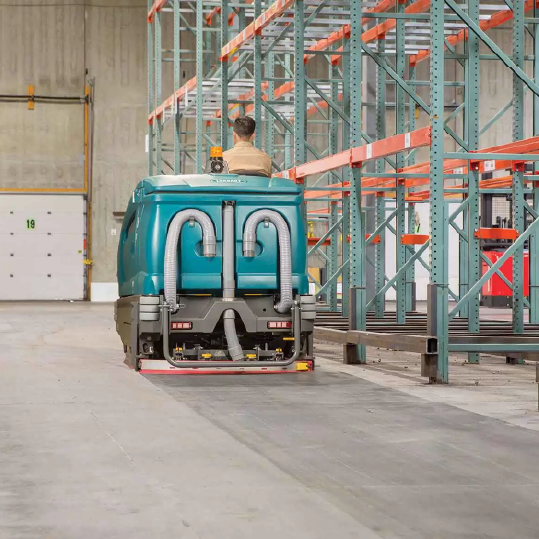 cleaning technician driving tennant rider floor scrubber down aisle in distribution center